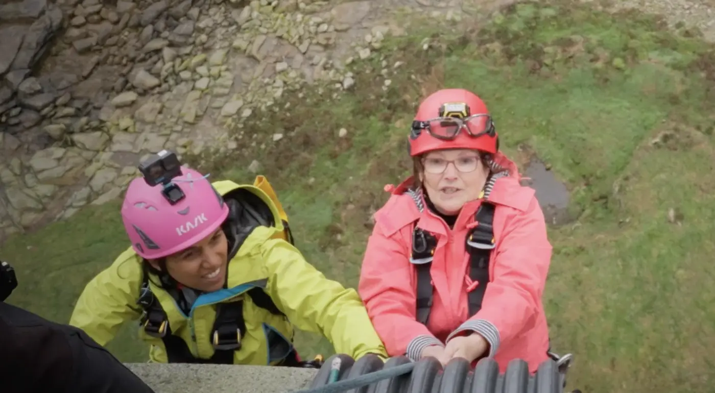marathon du vertige Deux challengeuses souriantes, l'une en casque rose, l'autre en casque rouge, se hissent sur un rocher herbeux.
