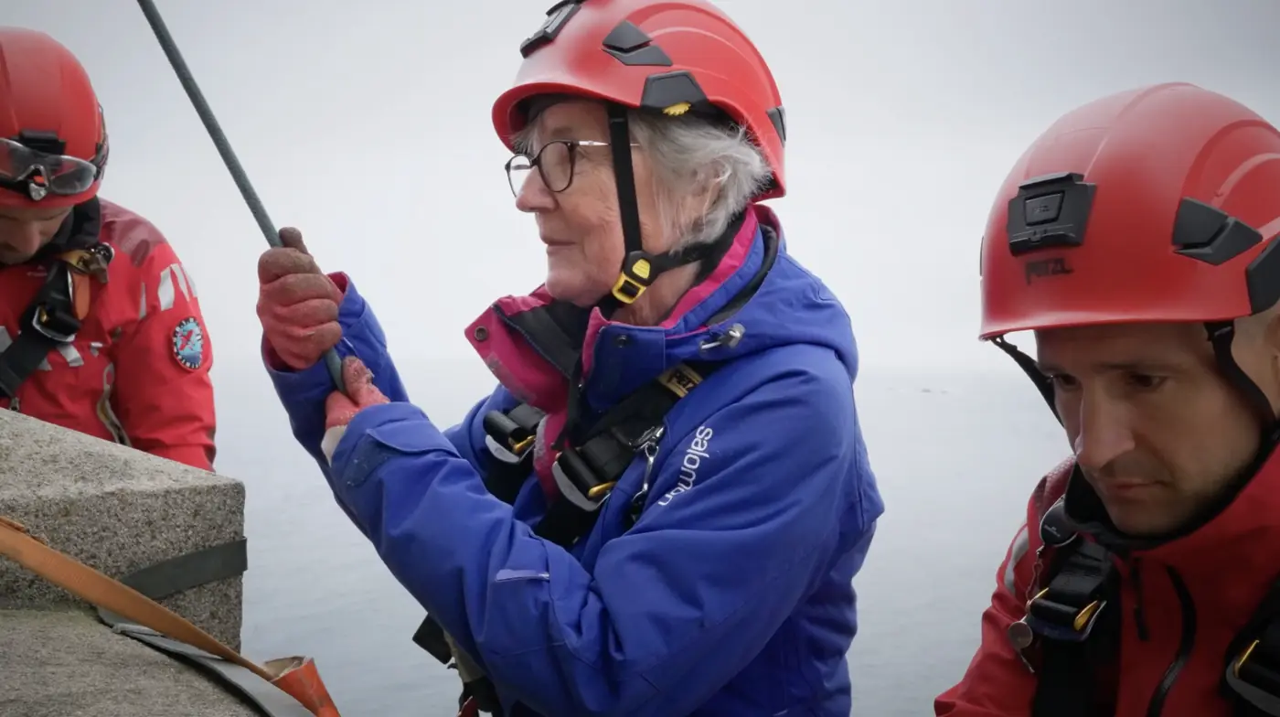 marathon du vertige Une challengeuse concentrée, casque rouge et veste bleue, sur le phare de l'Île Vierge, à côté d'un secouriste en tenue rouge.